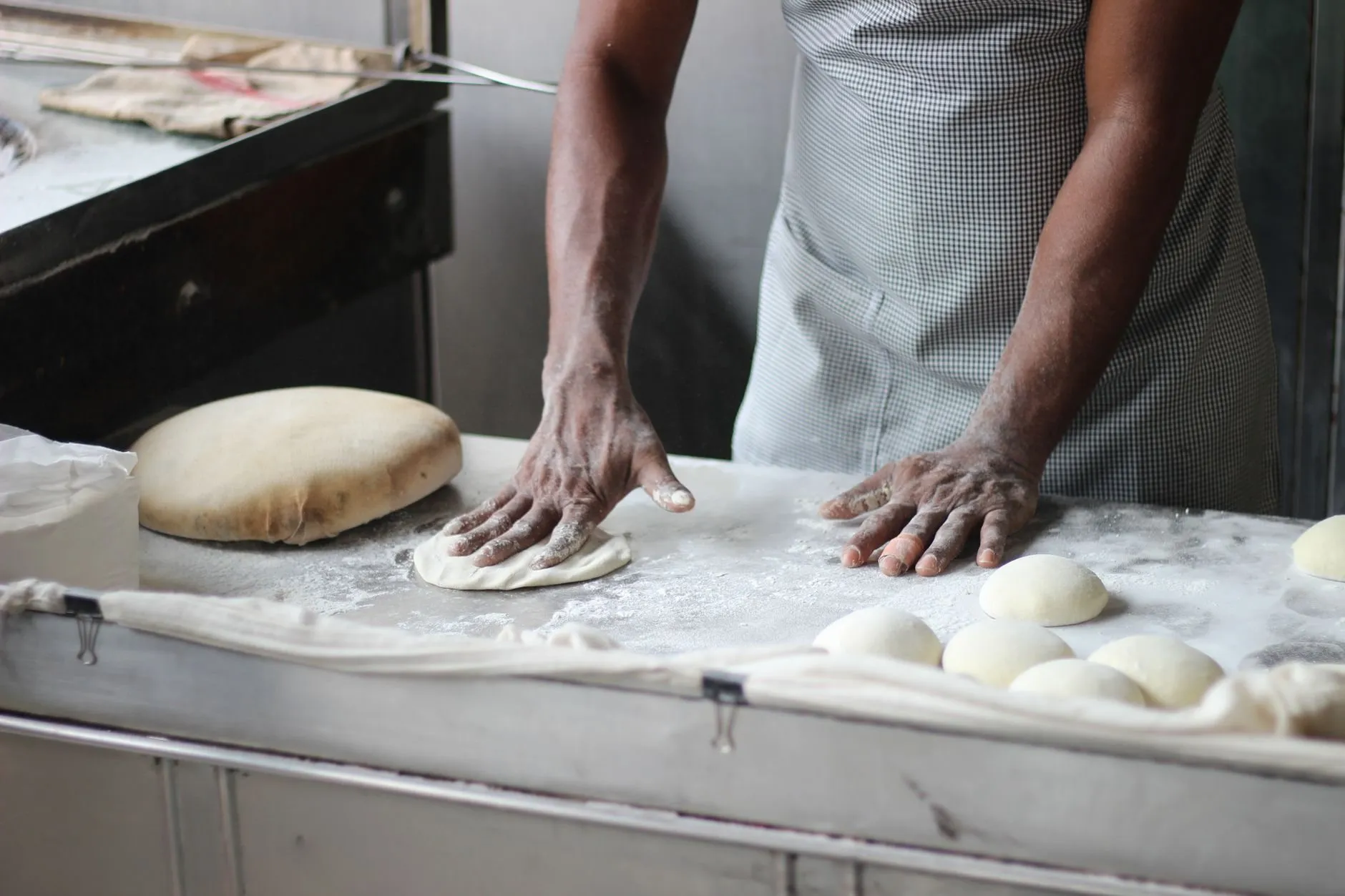 A baker's hands shaping dough on a floured steel bench
