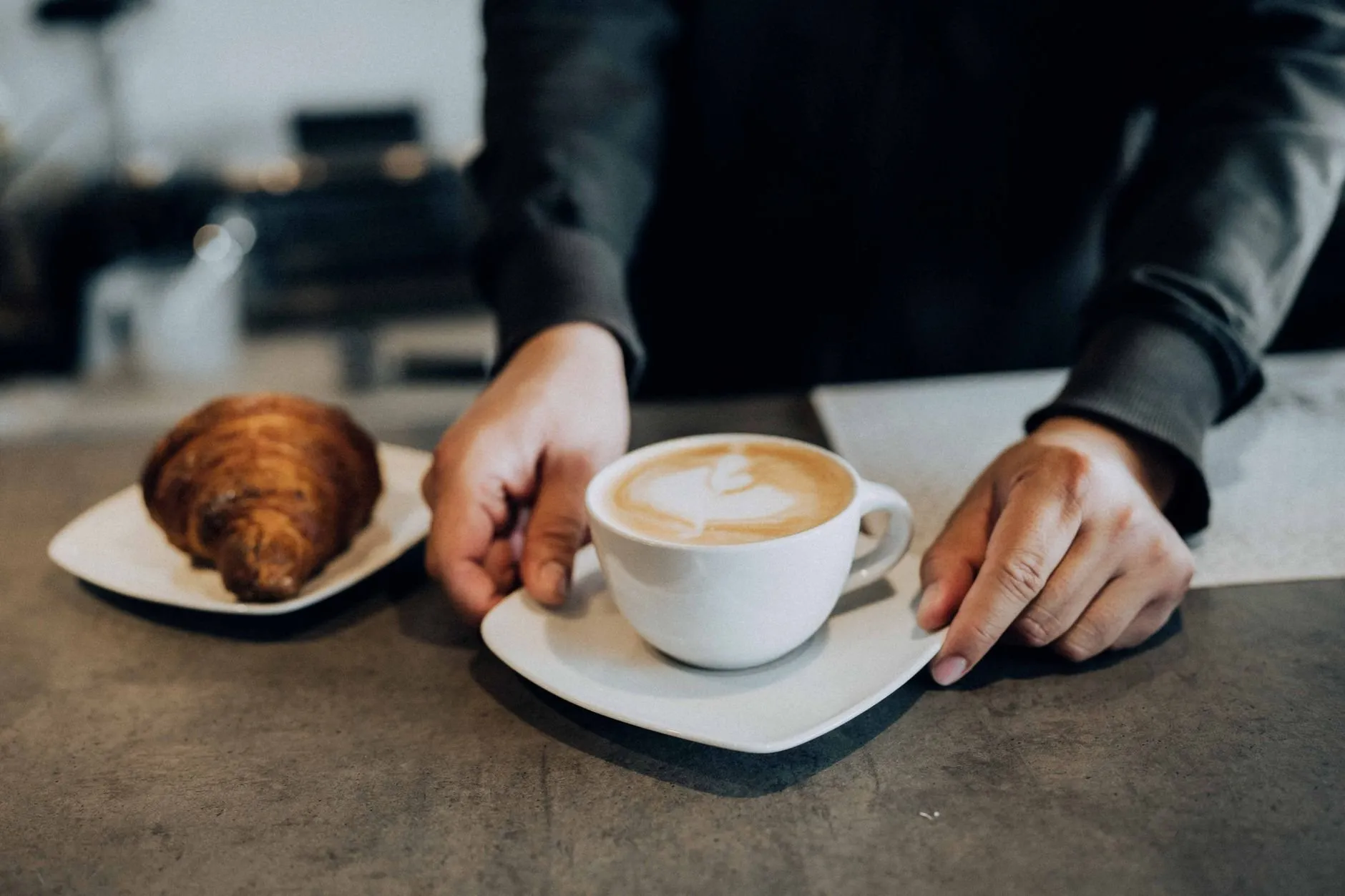 Hands setting down a latte beside a croissant on a café counter