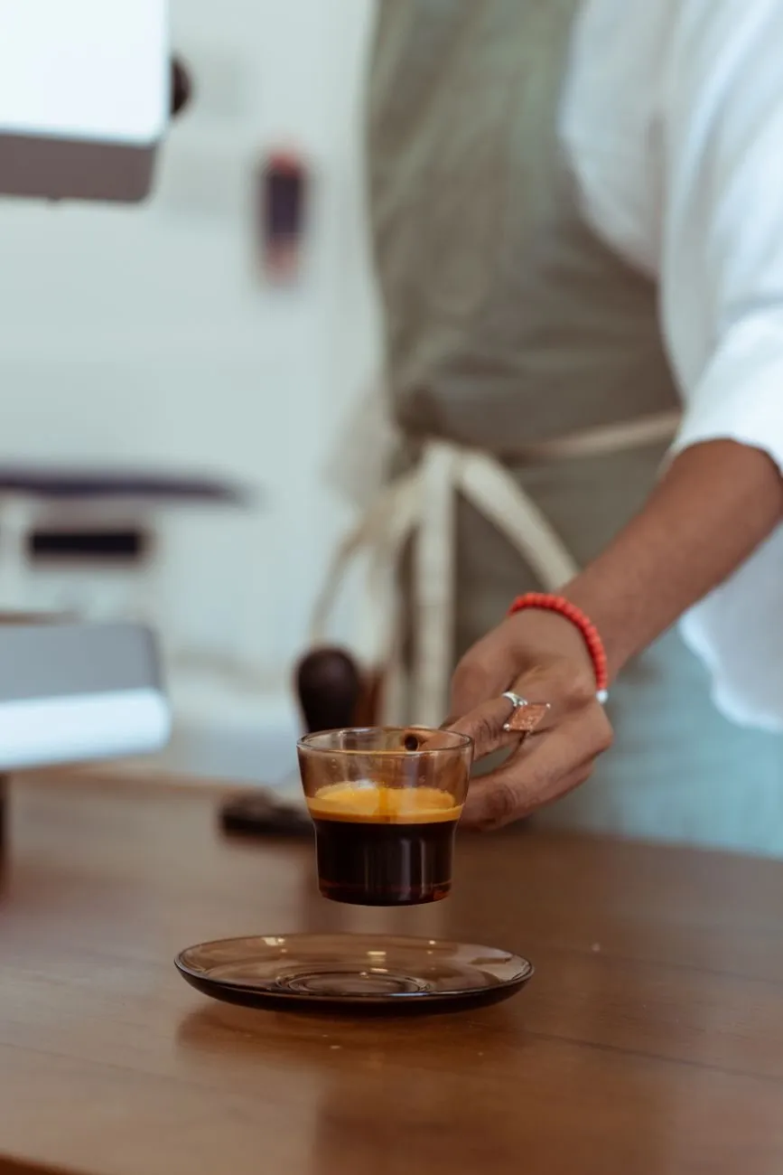 A barista's hand setting down a glass of espresso on a wooden counter