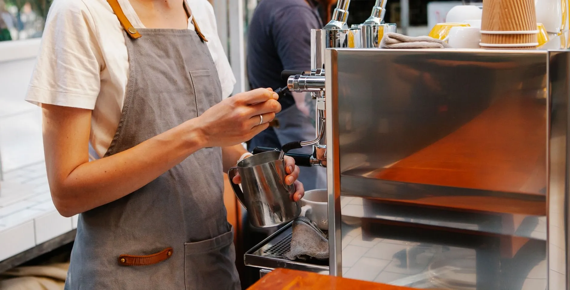 A barista in a grey apron steaming milk beside a steel espresso station