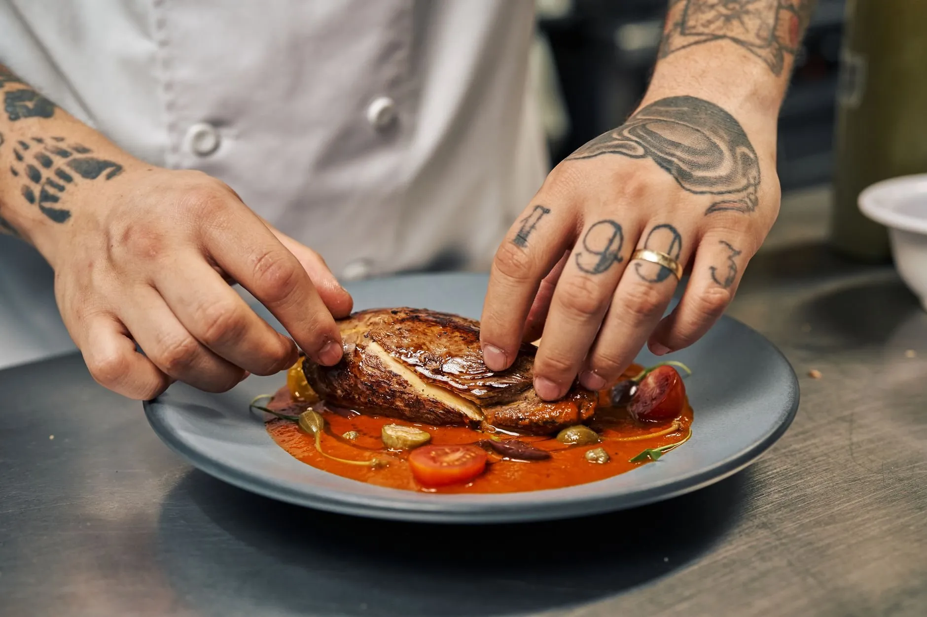 A chef's tattooed hands finishing a plated dish at the pass